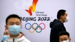 Participants wearing face masks gather near a logo for the 2022 Winter Olympic torch relay during an event to display the Olympic torch and flame at the Beijing University of Posts and Communications in Beijing, Thursday, Dec. 9, 2021. China is limiting the torch relay for the Winter Olympic Games to just three days amid coronavirus worries, organizers said Friday, Jan. 21, 2022. The flame will be displayed only in enclosed venues that are deemed "safe and controllable," according to officials speaking at a news conference. (AP Photo/Mark Schiefelbein)