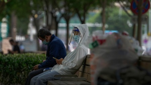 A woman wearing a face shield and mask takes a rest with other people on a green space Monday, May 23, 2022, in Beijing. Beijing extended orders for workers and students to stay home and ordered additional mass testing Monday as cases of COVID-19 again rose in the Chinese capital. (AP Photo/Andy Wong)