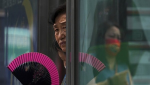 A woman fans herself at an advertisement boards on a bus stand reflecting a masked commuter as they wait for buses in Beijing, Tuesday, July 5, 2022. (AP Photo/Andy Wong)