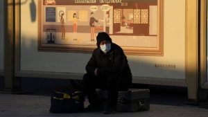 A man wearing a mask sits on his luggage near a mural depicting health checks before entry to a restaurant in Beijing, Thursday, Dec. 8, 2022. A day after China announced the rollback of some of its most stringent COVID-19 restrictions, people across the country are greeting the news with a measure of relief but also caution, as many wait to see how the new approach will be implemented. (AP Photo/Ng Han Guan)
