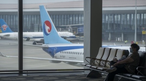 A traveler wearing a face mask sits near a parked jetliner from Hebei Airlines at Beijing Daxing International Airport in Beijing, Sunday, May 28, 2023. China's first domestically made passenger jet flew its maiden commercial flight on Sunday, as China looks to compete with industry giants such as Boeing and Airbus in the global aircraft market. (AP Photo/Mark Schiefelbein)