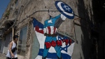 A woman walks past a mural depicting U.S. President Joe Biden as a superhero defending Israel on a street in Tel Aviv, Israel, Sunday, April 14, 2024. Israel on Sunday hailed its successful air defenses in the face of an unprecedented attack by Iran, saying it and its allies thwarted 99 per cent of the more than 300 drones and missiles launched toward its territory. But regional tensions remain high, amid fears of further escalation in the event of a possible Israeli counter-strike. (AP Photo/Leo Correa)
