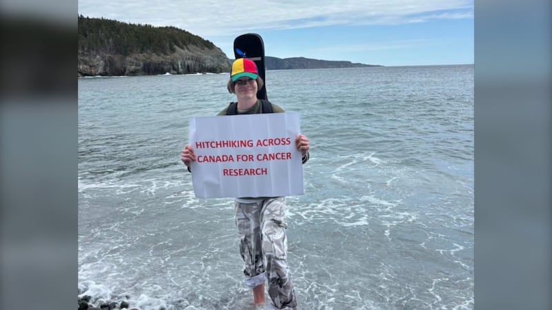 A young man stands in the ocean holding a sign that reads: "Hitchhiking across Canada for Cancer Research."