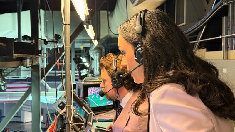 A woman with brown hair is wearing a headset microphone and a pink suit in a broadcasting booth.