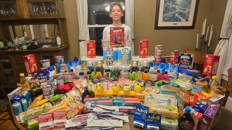 A teenage boy stands behind a kitchen table which is covered in donations for a food bank.