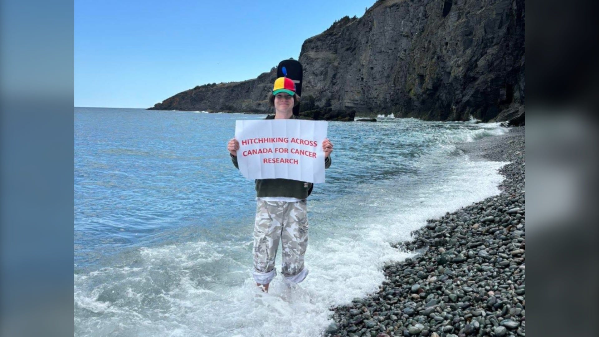 A young man stands in the ocean holding a sign that reads: "Hitchhiking across Canada for Cancer Research."
