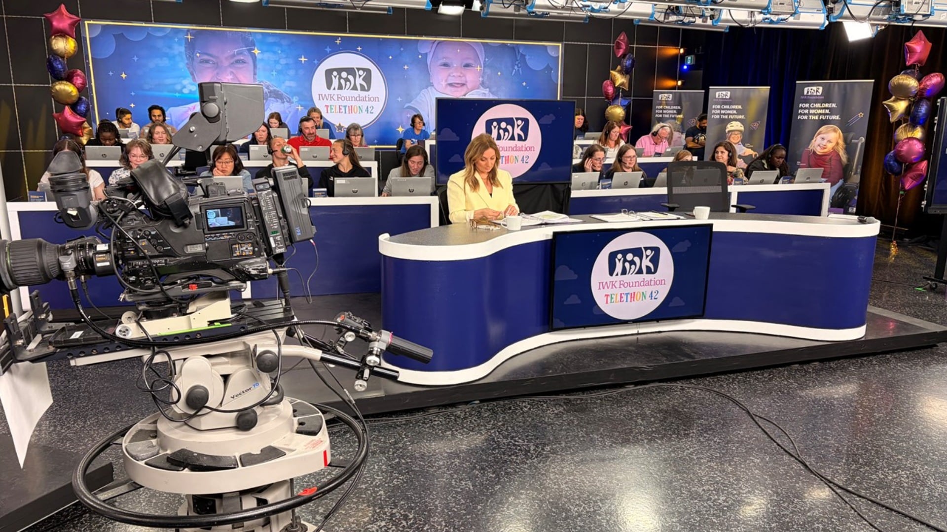 A woman wearing a yellow blazer sits at a news desk with people on computers behind her.
