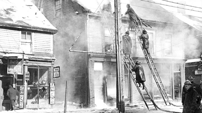A black and white photo shows firefighters climbing a ladder to battle a fire.