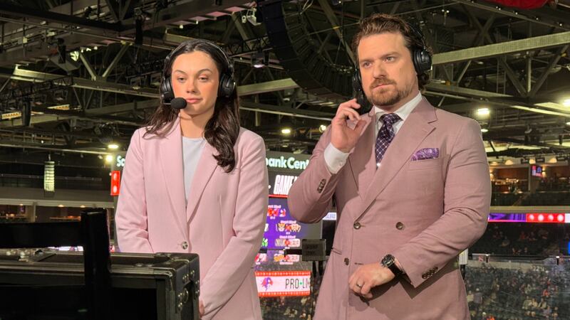 A woman in a pink suit stands next to a man in a mauve suit during a television broadcast.